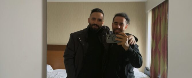 Two men of Syrian heritage stand next to each other taking a selfie in a mirror. One has his arm around the shoulder of the other in a relaxed and friendly way, and they are smiling.