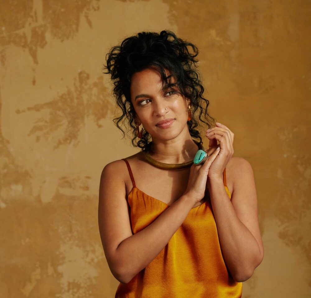 A woman with dark wavey hair and an orange dress is smiling and clasping her hands together