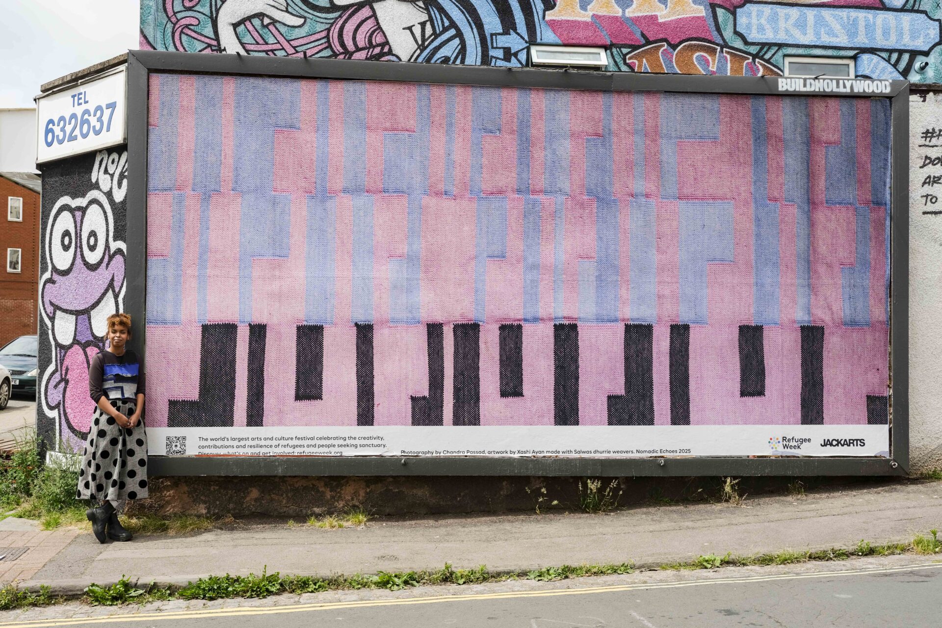 Ayan stands next to a large billboard on the street fearuring a detail of a woven piece