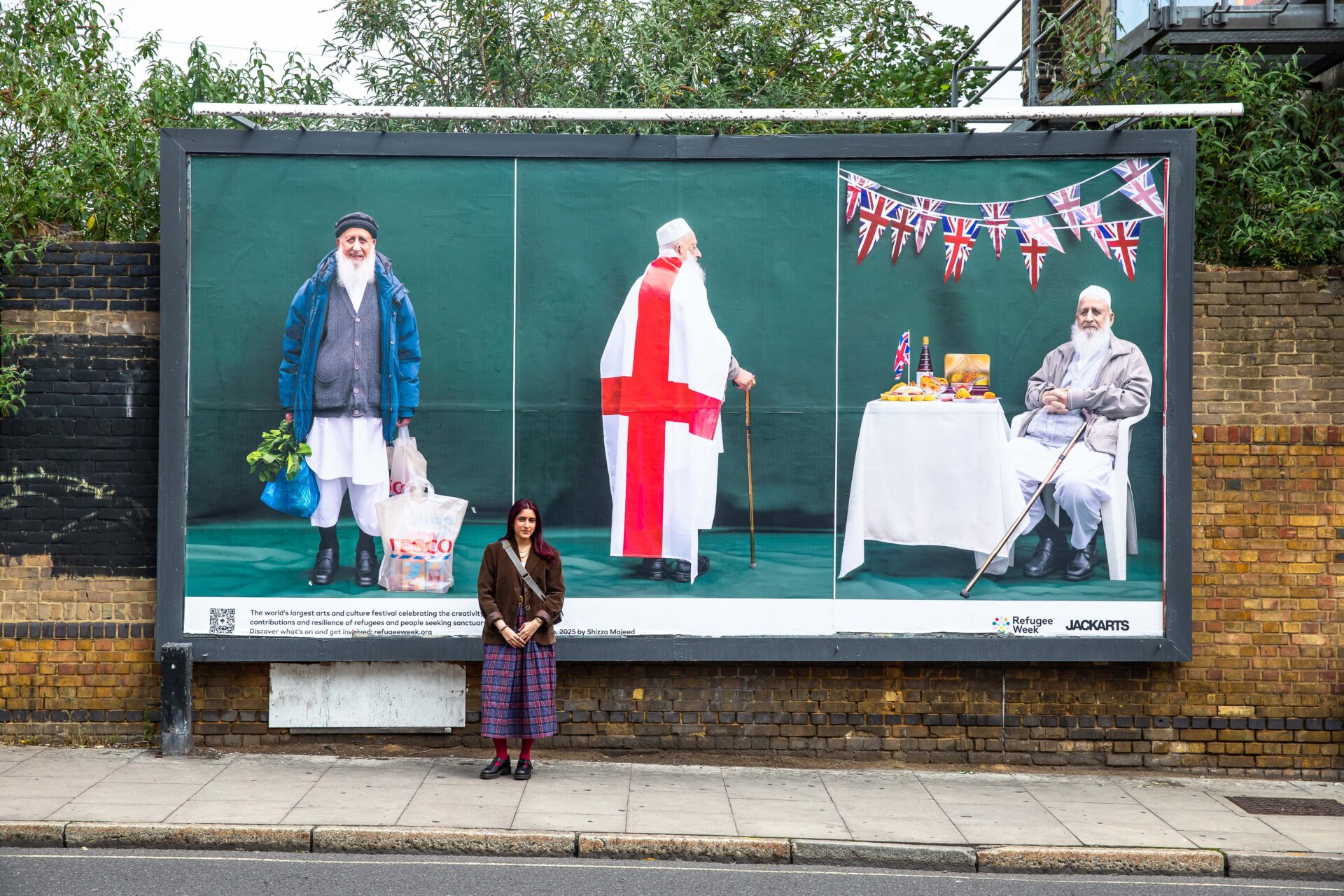A billboard on the street featuring three portraits of a pakistani man in traditionalk dress draped in the george cross flag, with union jack bunting and with a tesco's shopping bag. The artist stands in front of the billboard.