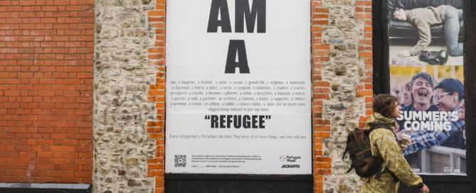 A man walk past a large poster billboard on the street - it says I AM A REFUGEE in large bold letters, but also goes on to list all the other things i am including a daughterm a brother, a friends, a classmate, a thinker, a runner and more...